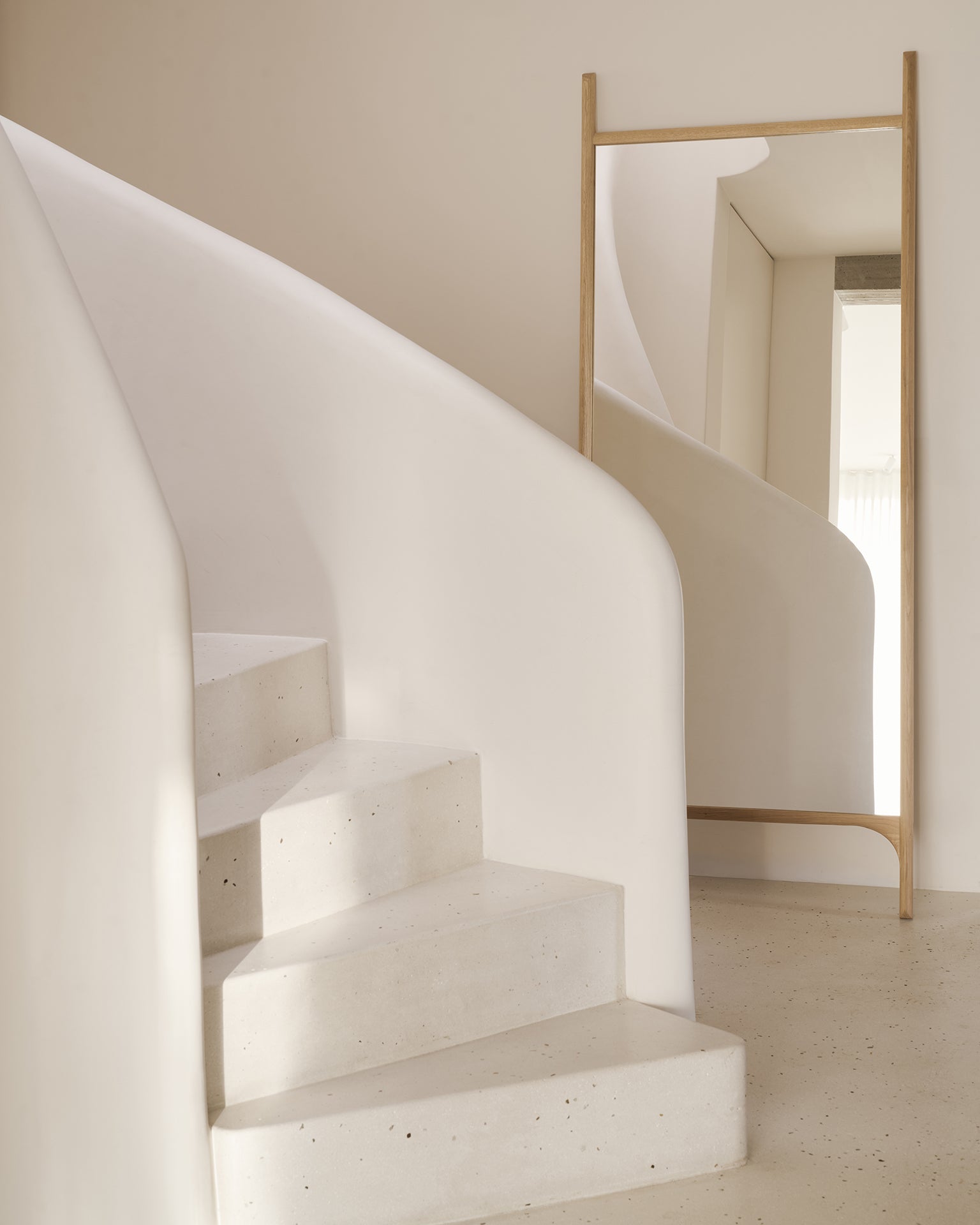 A minimalist interior with a white curved staircase sits beside Ethnicraft’s PI Floor Mirror – Oak, its solid oak frame and polished imperfect finish reflecting the stairs and enhancing the clean, modern look with smooth surfaces and neutral tones.
