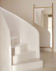 A minimalist interior with a white curved staircase sits beside Ethnicraft’s PI Floor Mirror – Oak, its solid oak frame and polished imperfect finish reflecting the stairs and enhancing the clean, modern look with smooth surfaces and neutral tones.