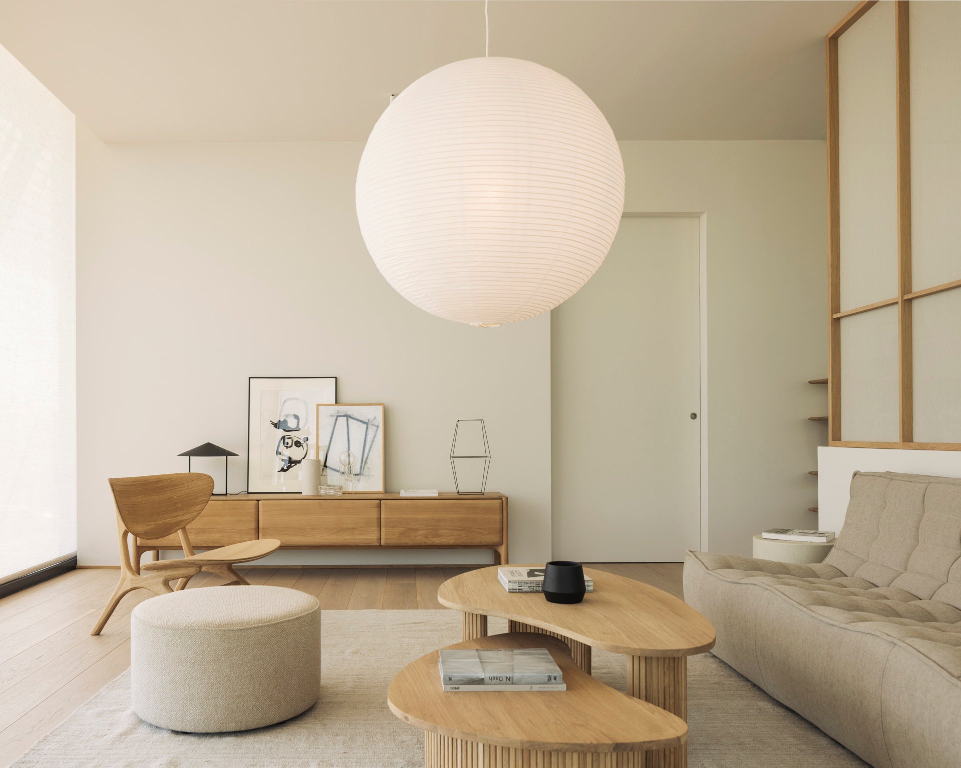 A minimalist living room in neutral tones features a large round paper lantern and light wood furniture, including the Ethnicraft PI TV Cupboard crafted from solid oak. A low sofa faces framed art displayed on a sideboard, with a round ottoman placed on a rug. Sunlight pours in through a large window on the left.