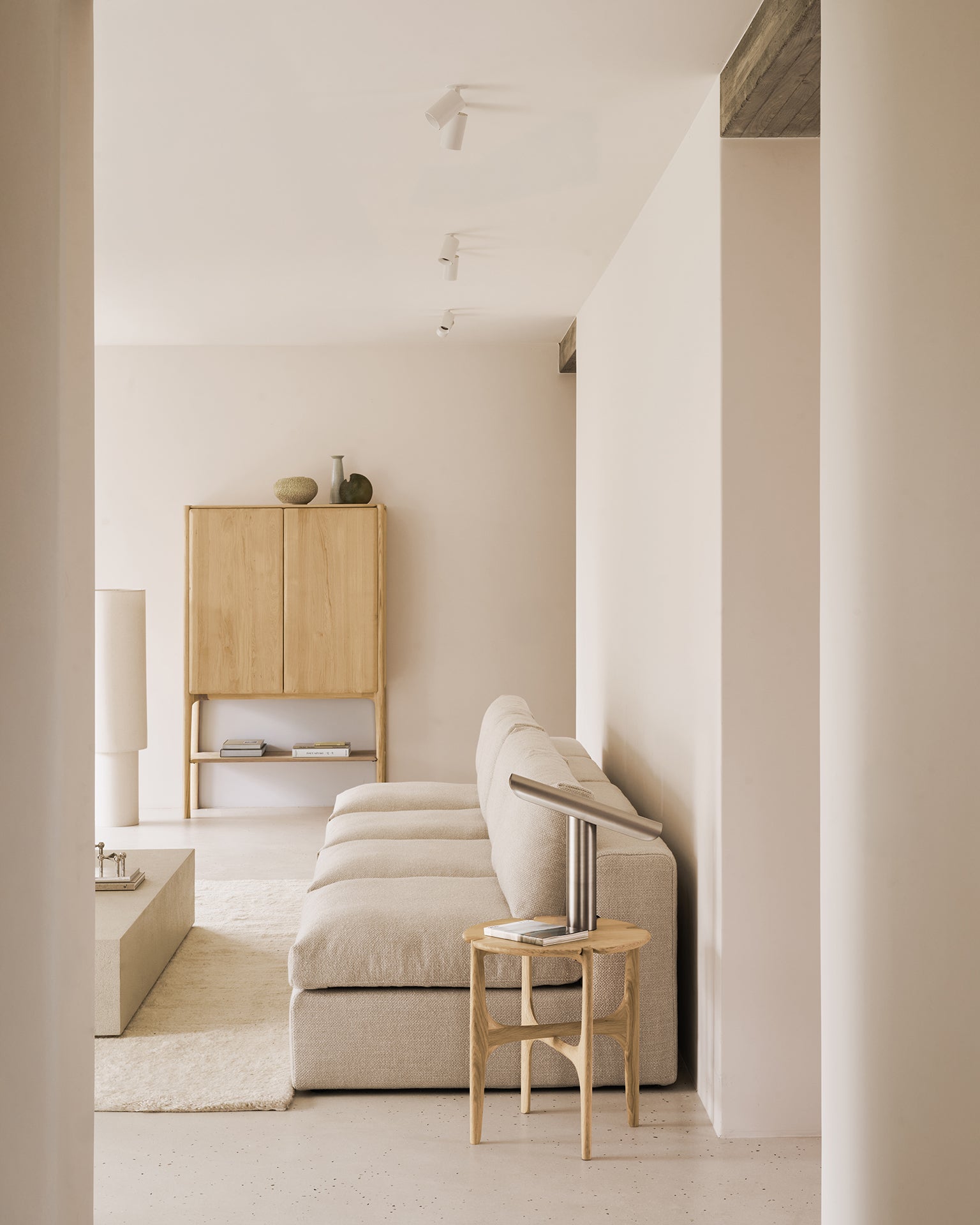 A minimalist living room with neutral tones features a light beige sectional sofa, light wood side table, square coffee table, and the Ethnicraft PI Cupboard - Oak as handcrafted storage, all set against cream-colored walls and floor.
