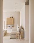 A minimalist living room with neutral tones features a light beige sectional sofa, light wood side table, square coffee table, and the Ethnicraft PI Cupboard - Oak as handcrafted storage, all set against cream-colored walls and floor.