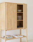 The Ethnicraft PI Cupboard in oak stands against a plain wall, its door ajar to show shelves with plates, glasses, and small containers. Two stacks of books rest on the lower shelf.