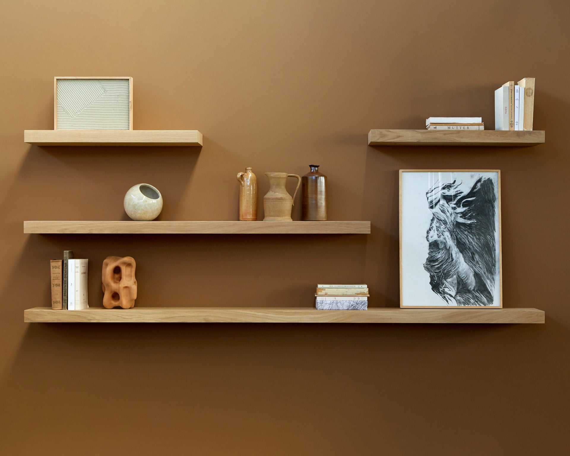 Three Ethnicraft Wall Shelf - Oak units are mounted on a brown wall, displaying books, ceramic vases, abstract art, and decorative items in a minimalist style.