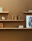 Three Ethnicraft Wall Shelf - Oak units are mounted on a brown wall, displaying books, ceramic vases, abstract art, and decorative items in a minimalist style.