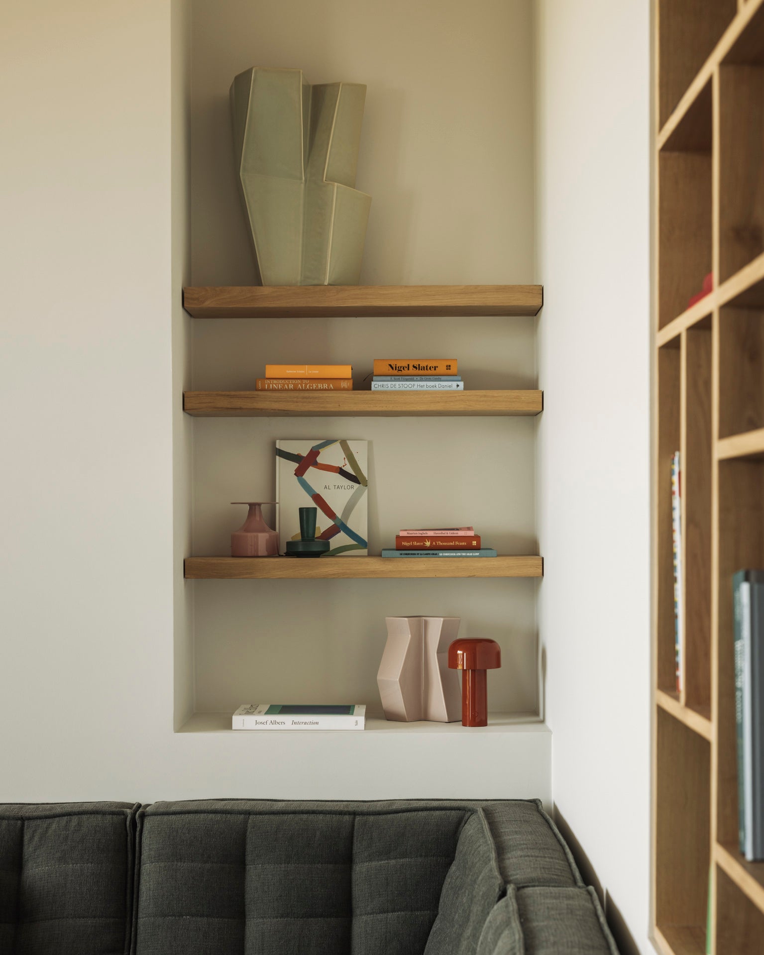 A modern living room features an Ethnicraft Wall Shelf - Oak displaying books, abstract vases, and décor above a gray tufted sofa. Soft beige, brown, and muted accent tones create a cozy atmosphere with a stylish neutral palette.