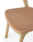 A close-up of the Ethnicraft Bok Dining Chair, made from solid oak with light wood legs and a sienna leather seat cushion, photographed against a white background.