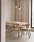 A minimalist dining area with Ethnicraft’s Bok Extendable Dining Table in varnished oak, four matching chairs, a white bowl, neutral curtains, a large window, and two modern pendant lights overhead.