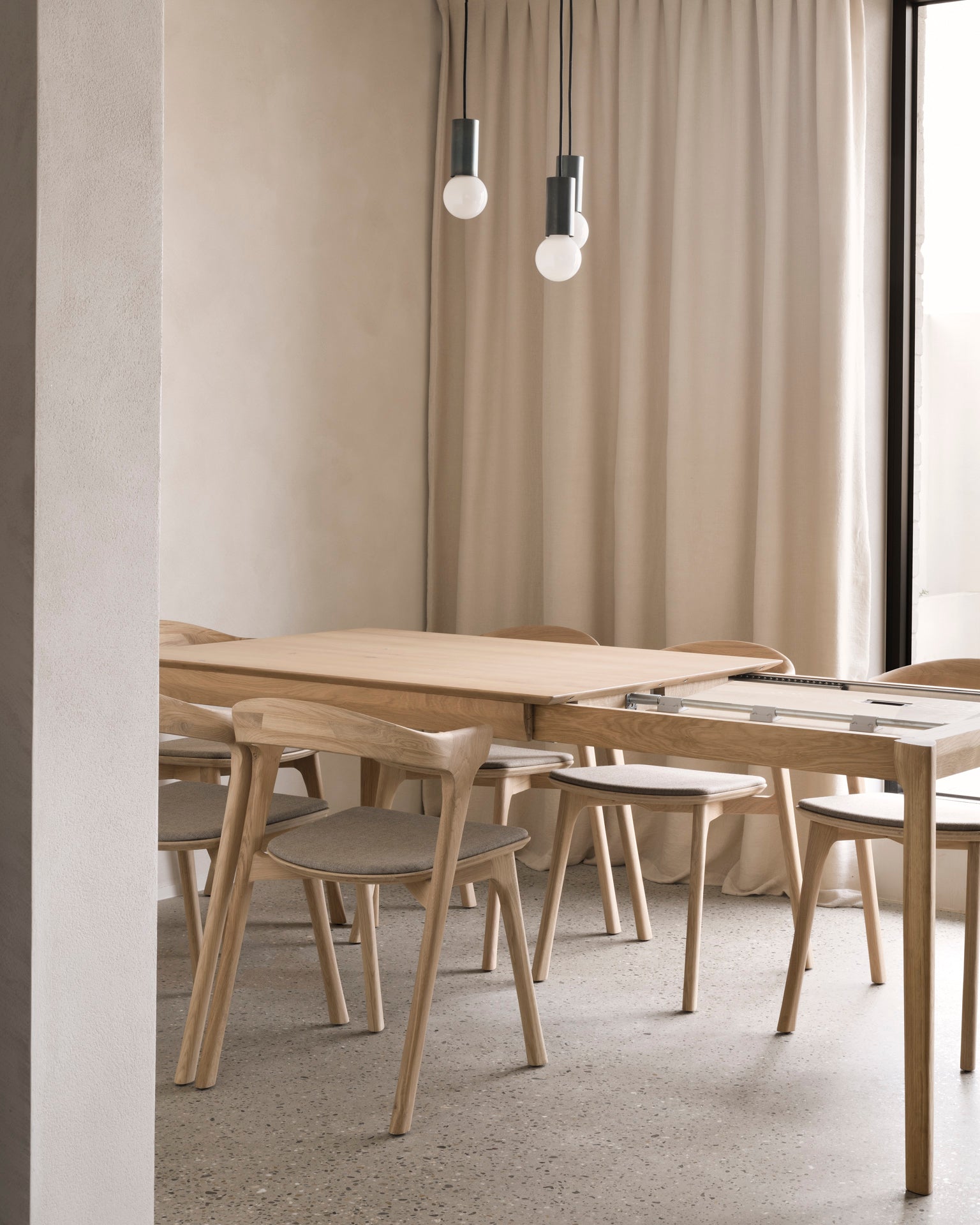 A minimalist dining area featuring the Ethnicraft Bok Extendable Dining Table in varnished oak, paired with light wood chairs, beige curtains, and modern pendant lights. The space showcases a soft, natural palette and clean lines.