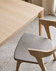 An Ethnicraft Bok Extendable Dining Table in varnished oak is paired with a matching chair featuring a curved backrest and cushioned beige seat, arranged on a speckled terrazzo floor beside beige curtains.