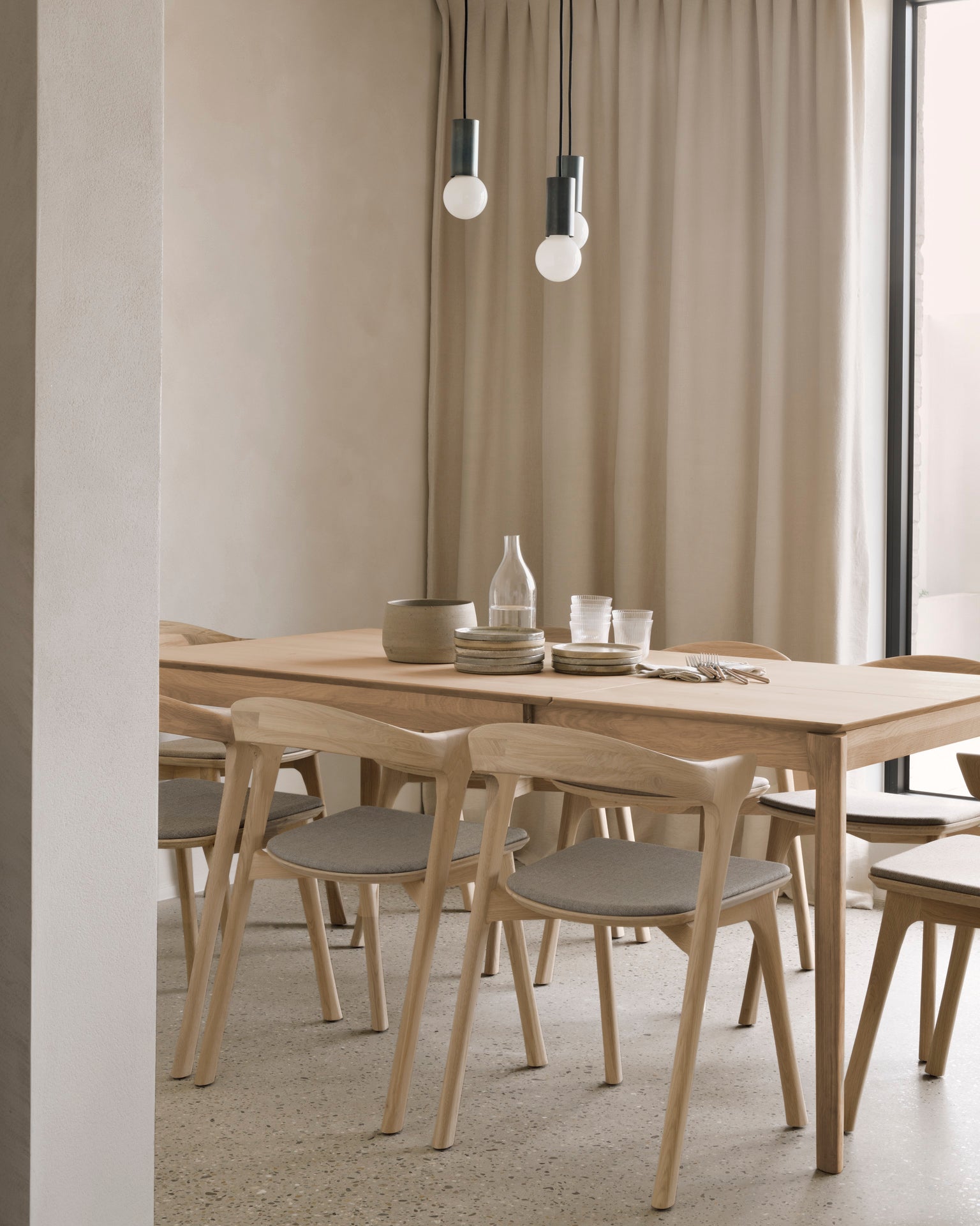 Minimalist dining room with an Ethnicraft Bok Extendable Dining Table in varnished oak, matching chairs, neutral dishes and glassware, beige curtains, terrazzo floor, and two pendant lights hanging from the ceiling.