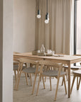 Minimalist dining room with an Ethnicraft Bok Extendable Dining Table in varnished oak, matching chairs, neutral dishes and glassware, beige curtains, terrazzo floor, and two pendant lights hanging from the ceiling.