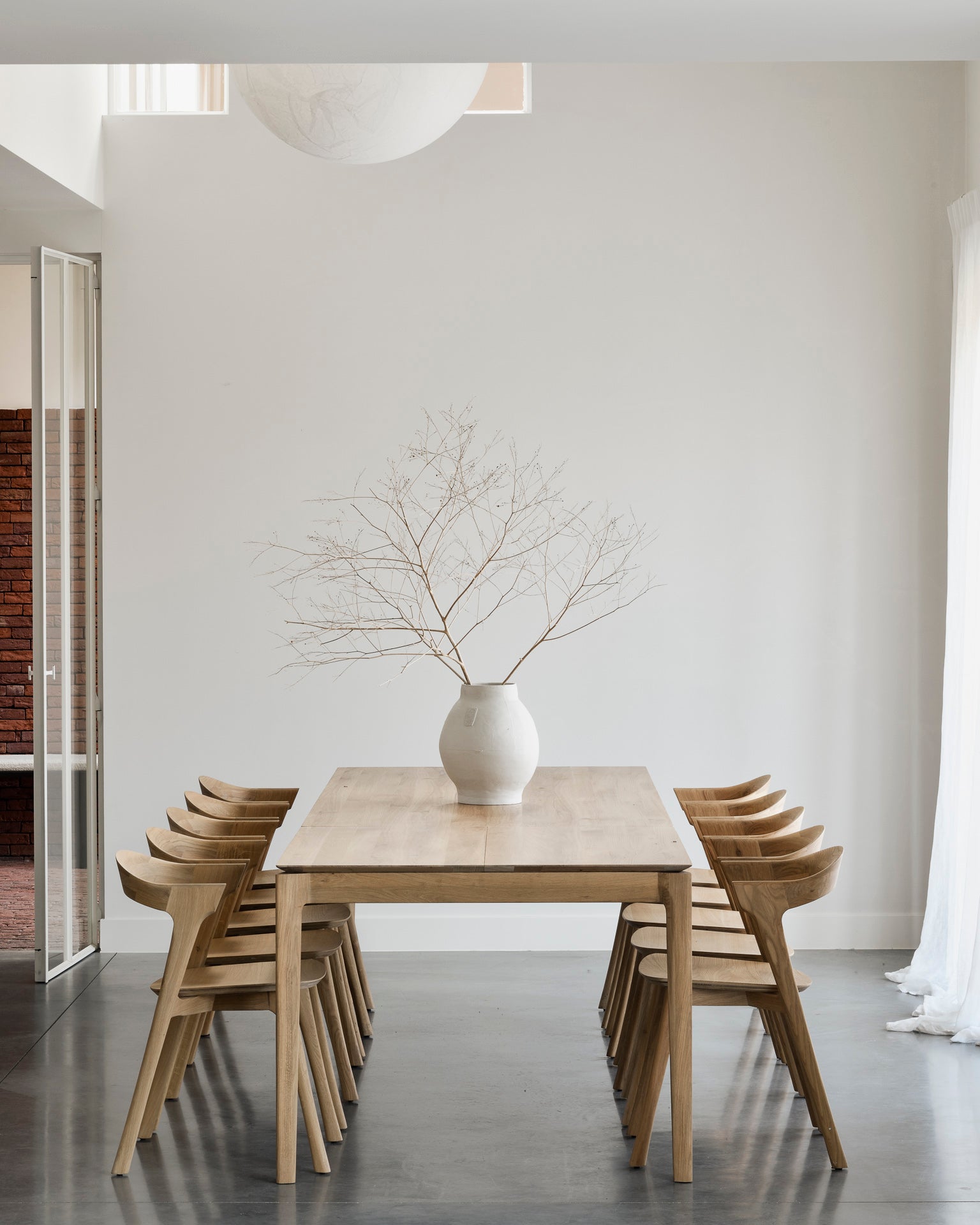A minimalist dining room features the Ethnicraft Bok Extendable Dining Table in oak, paired with eight wooden chairs and a white vase with sparse branches. White walls, high ceilings, and a large window fill the space with natural light.
