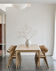 A minimalist dining room features the Ethnicraft Bok Extendable Dining Table in oak, paired with eight wooden chairs and a white vase with sparse branches. White walls, high ceilings, and a large window fill the space with natural light.