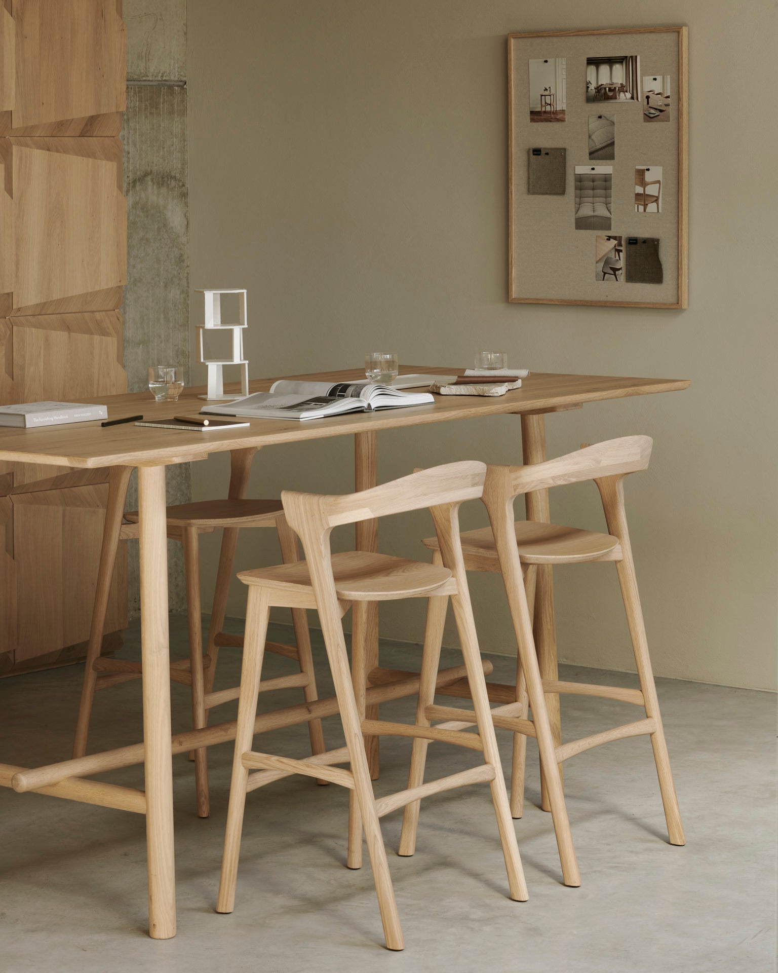 A minimalist dining space features two Ethnicraft Bok bar stools in oak at a tall table, with open books, glasses, and stationery on top. A corkboard with photos and notes decorates the beige wall in the background.