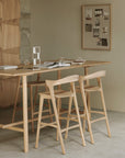 A minimalist dining space features two Ethnicraft Bok bar stools in oak at a tall table, with open books, glasses, and stationery on top. A corkboard with photos and notes decorates the beige wall in the background.