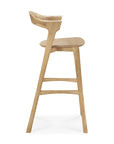 Side view of the Ethnicraft Bok bar stool - Oak, featuring a curved backrest, gently angled legs, a footrest, and a natural solid oak finish.