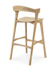 The Ethnicraft Bok bar stool in oak features a contoured seat, curved backrest, angled legs, and sturdy footrests. Crafted from solid oak with a simple natural finish, it combines modern design with comfort and support.