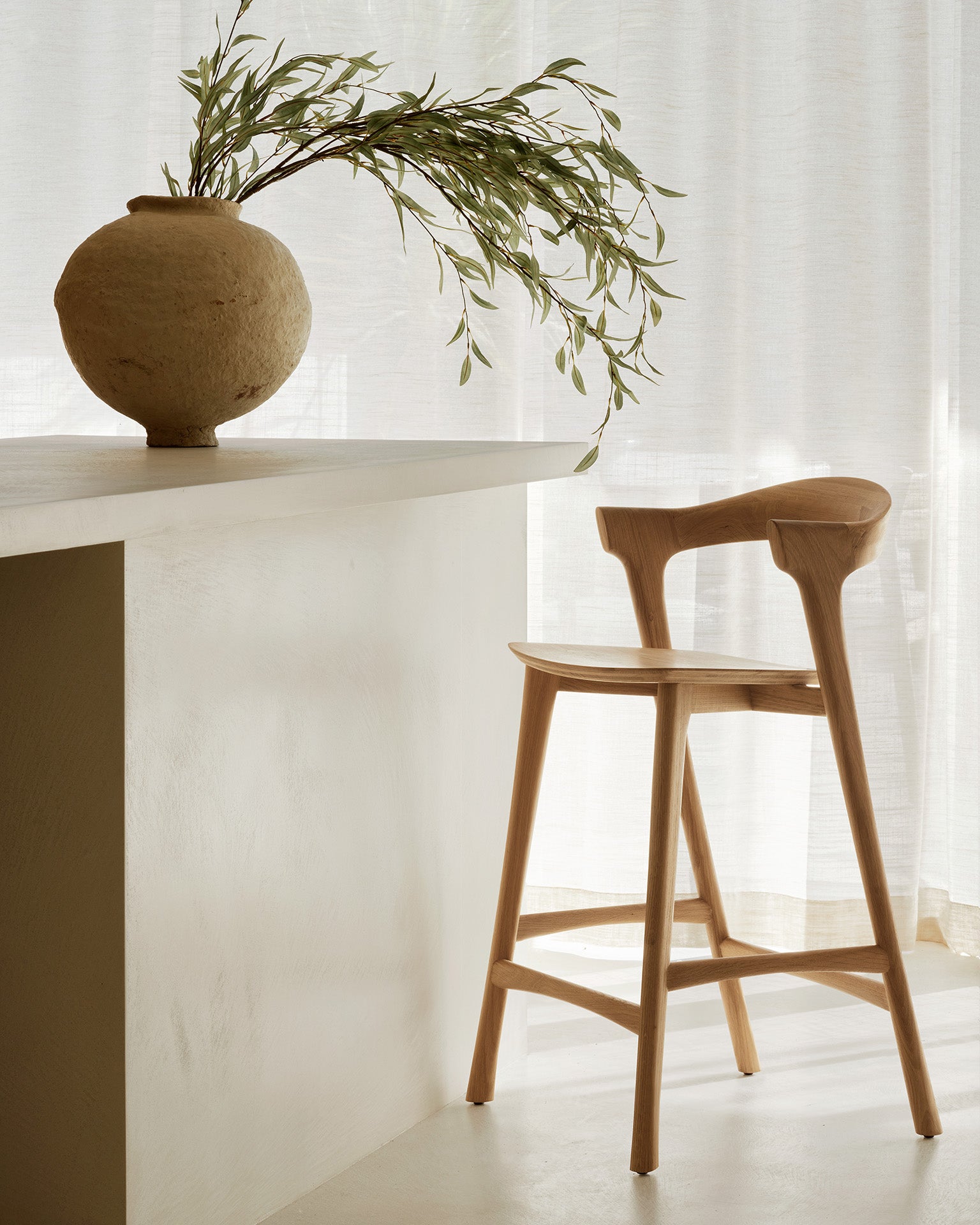 An Ethnicraft Bok Counter Stool in solid oak sits by a white counter with a textured vase of green branches, accented by sheer curtains in a sunlit, minimalist room.
