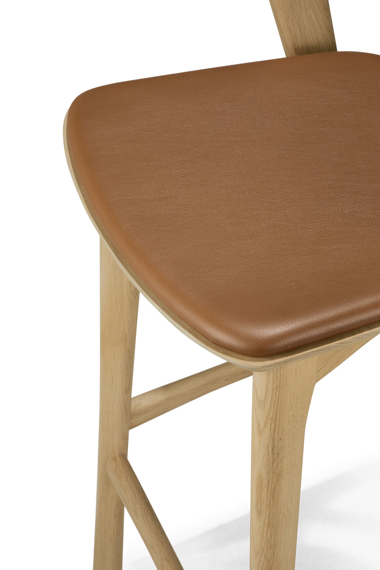 A close-up of the Bok bar stool by Ethnicraft, showcasing smooth solid oak legs and a light brown leather-upholstered seat. The minimalist design emphasizes the natural beauty of the wood.