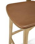 A close-up of the Bok bar stool by Ethnicraft, showcasing smooth solid oak legs and a light brown leather-upholstered seat. The minimalist design emphasizes the natural beauty of the wood.