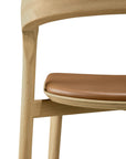 Close-up of the Ethnicraft Bok bar stool in solid oak, showcasing its curved backrest and tan leather seat cushion, set against a white background.
