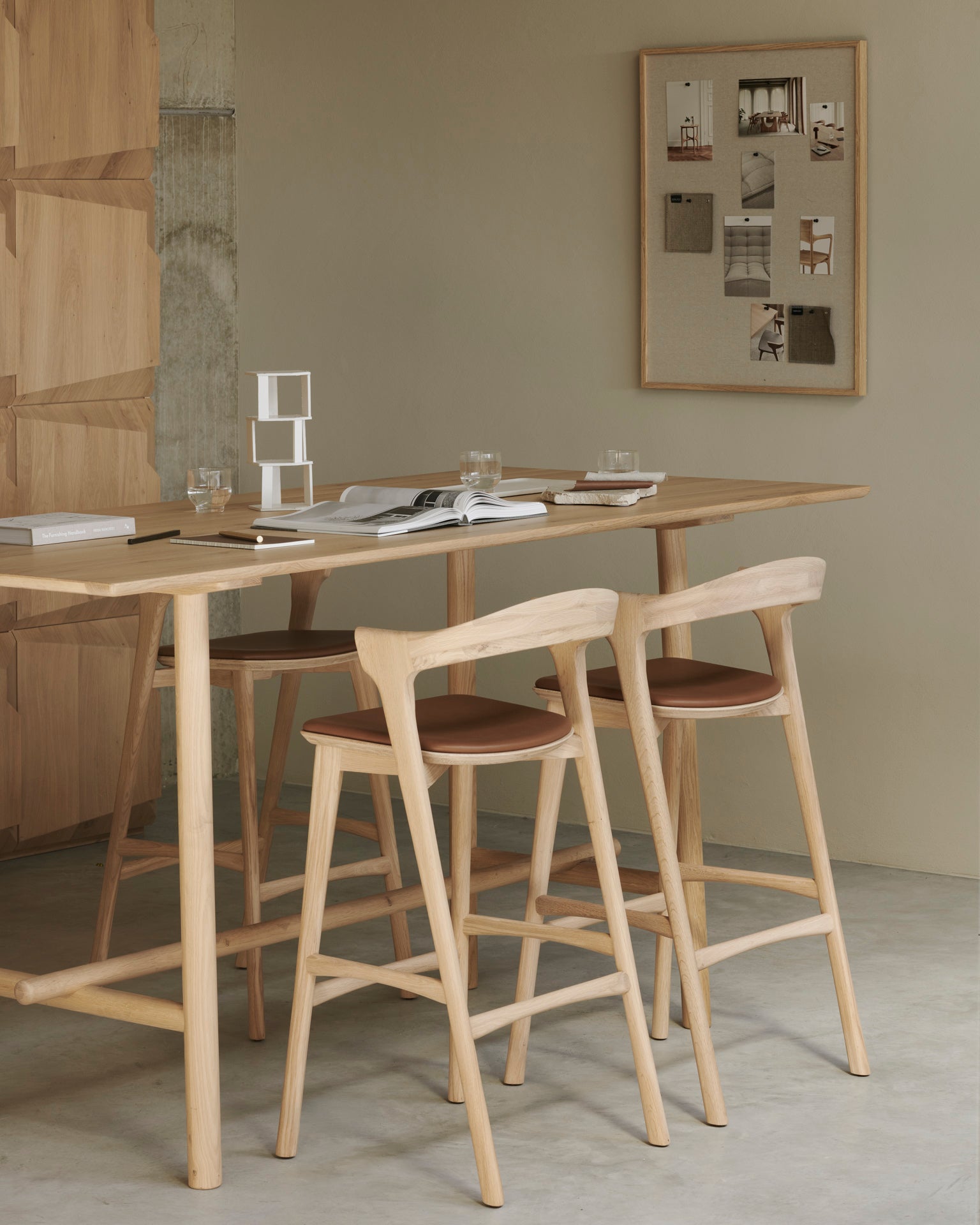 A minimalist dining area featuring a light solid oak table, two Ethnicraft Bok bar stools in oak, books and glasses on the table, and a bulletin board with photos on a beige wall in the background.