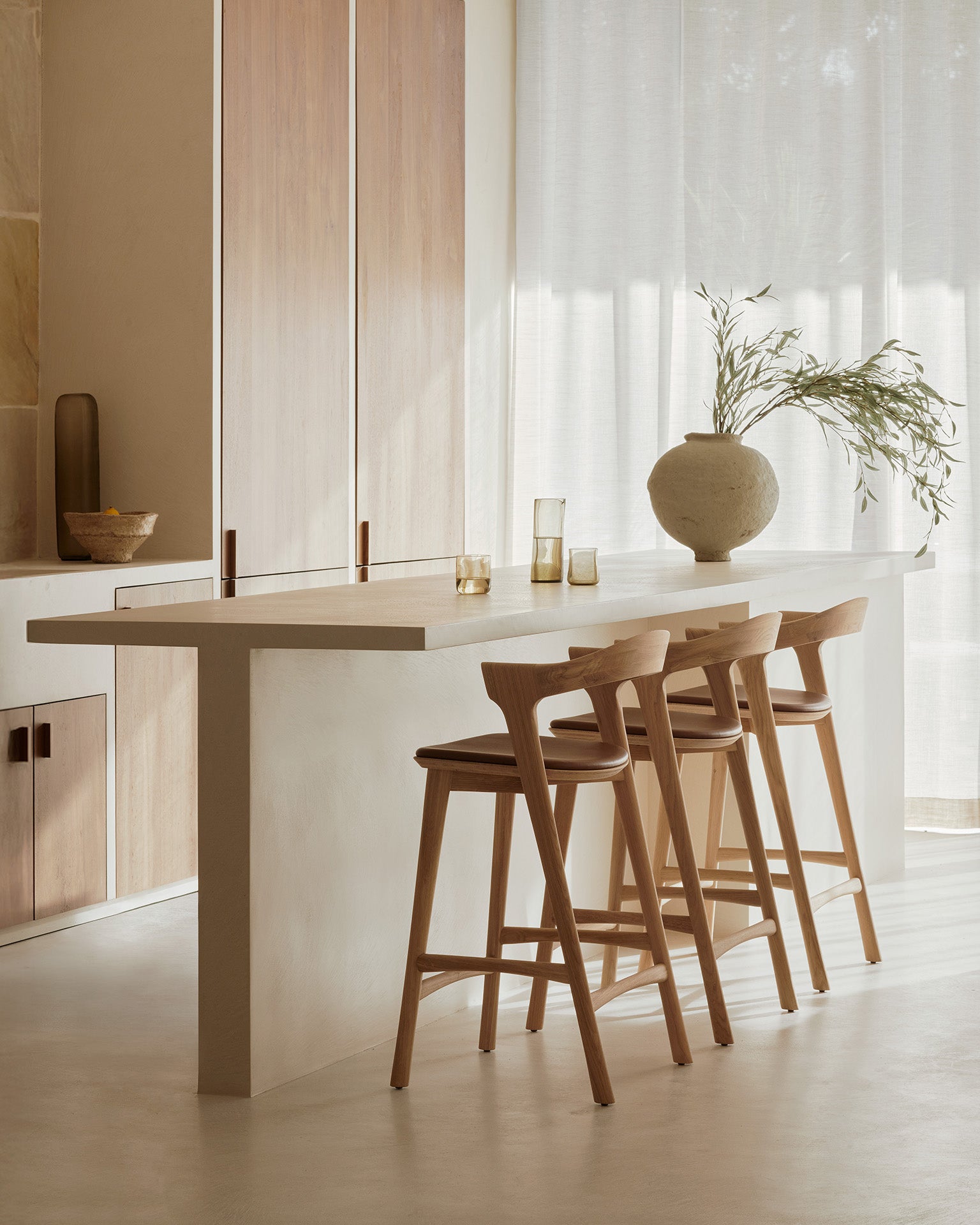 A minimalist kitchen features a white island, three Ethnicraft Bok bar stools in solid oak, a large ceramic vase with green branches, glass tumblers, and soft natural light through sheer curtains for a serene, neutral-toned atmosphere.