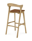The Bok bar stool - Oak by Ethnicraft features a solid oak frame, angled legs, curved backrest, and a modern open design with a brown cushioned seat.