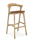 The Ethnicraft Bok bar stool - Oak features a modern design with a curved backrest, solid oak frame, brown cushioned seat, and four angled legs linked by sturdy supports.