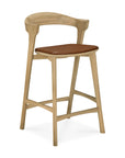 The Ethnicraft Bok counter stool features a light solid oak frame, curved backrest, footrest, and a brown cushioned seat inspired by the Bok dining chair.
