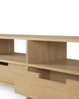 A light oak TV stand crafted by Ethnicraft, the Kabuki TV cupboard features open shelving and a partially open lower drawer, its minimalist silhouette displayed against a white background.