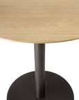 A close-up of the Ethnicraft Blend Dining Table - Oak shows its round solid oak top with a light finish and a single black metal pedestal base on a circular bottom, set against a white background.