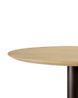 The Ethnicraft Blend Dining Table - Oak features a round solid oak top with a light finish, a single black cylindrical leg, and interchangeable tops, showcased against a plain white background.