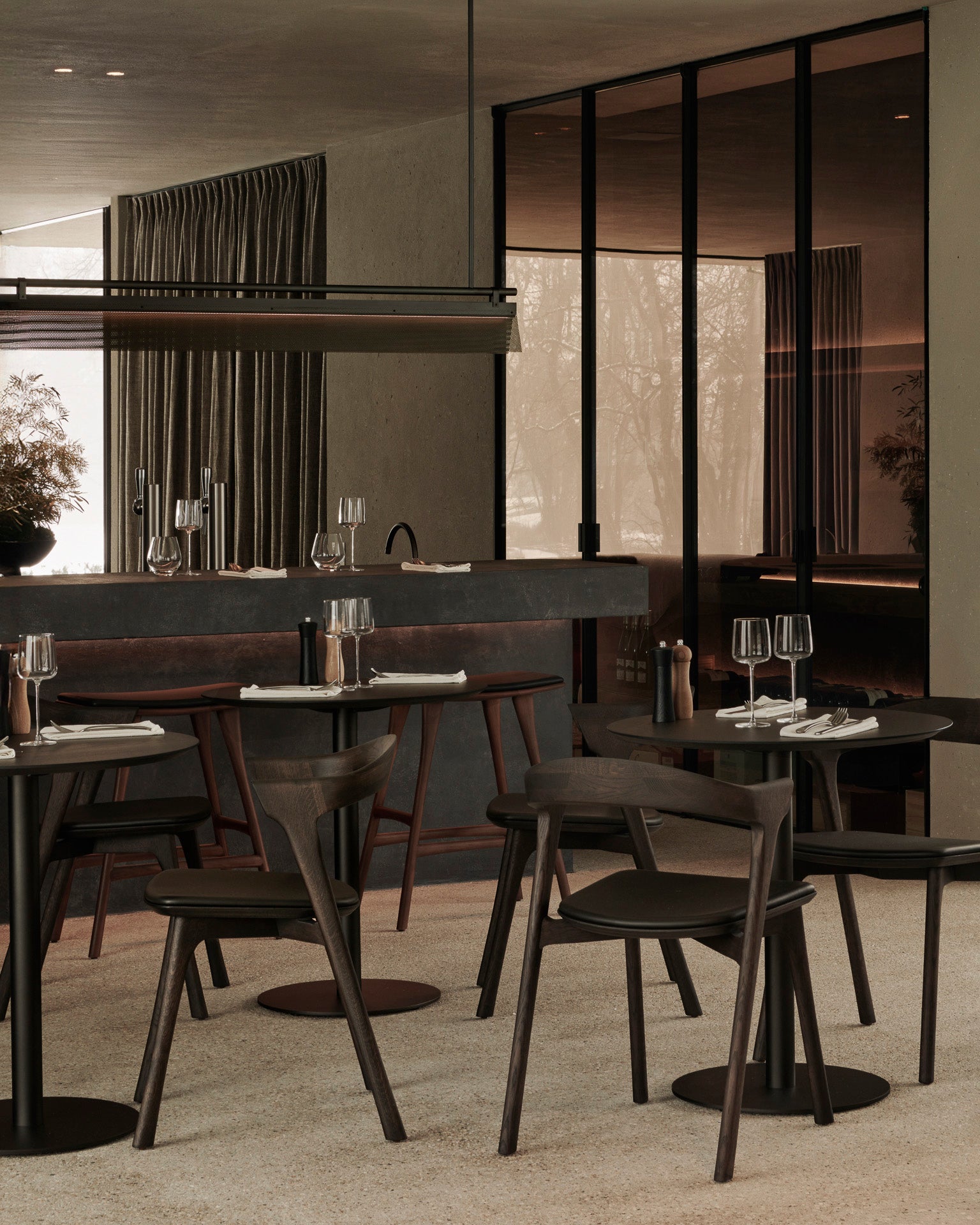 Modern restaurant interior featuring Ethnicraft’s Blend Dining Table in Brown Oak, paired with matching chairs. The space includes large windows, a sleek bar, minimalist décor, and table settings with wine glasses and napkins.