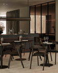 Modern restaurant interior featuring Ethnicraft’s Blend Dining Table in Brown Oak, paired with matching chairs. The space includes large windows, a sleek bar, minimalist décor, and table settings with wine glasses and napkins.