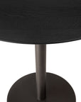 A close-up of the Ethnicraft Blend Dining Table - Black Oak highlights its round solid oak tabletop and metal pedestal base, set against a white background.