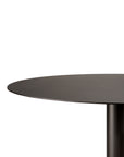 A close-up of the Ethnicraft Blend Dining Table - Dark Bronze Metal shows a round, dark bronze top with a single cylindrical leg. Against a white background, only part of the tabletop and leg are visible.