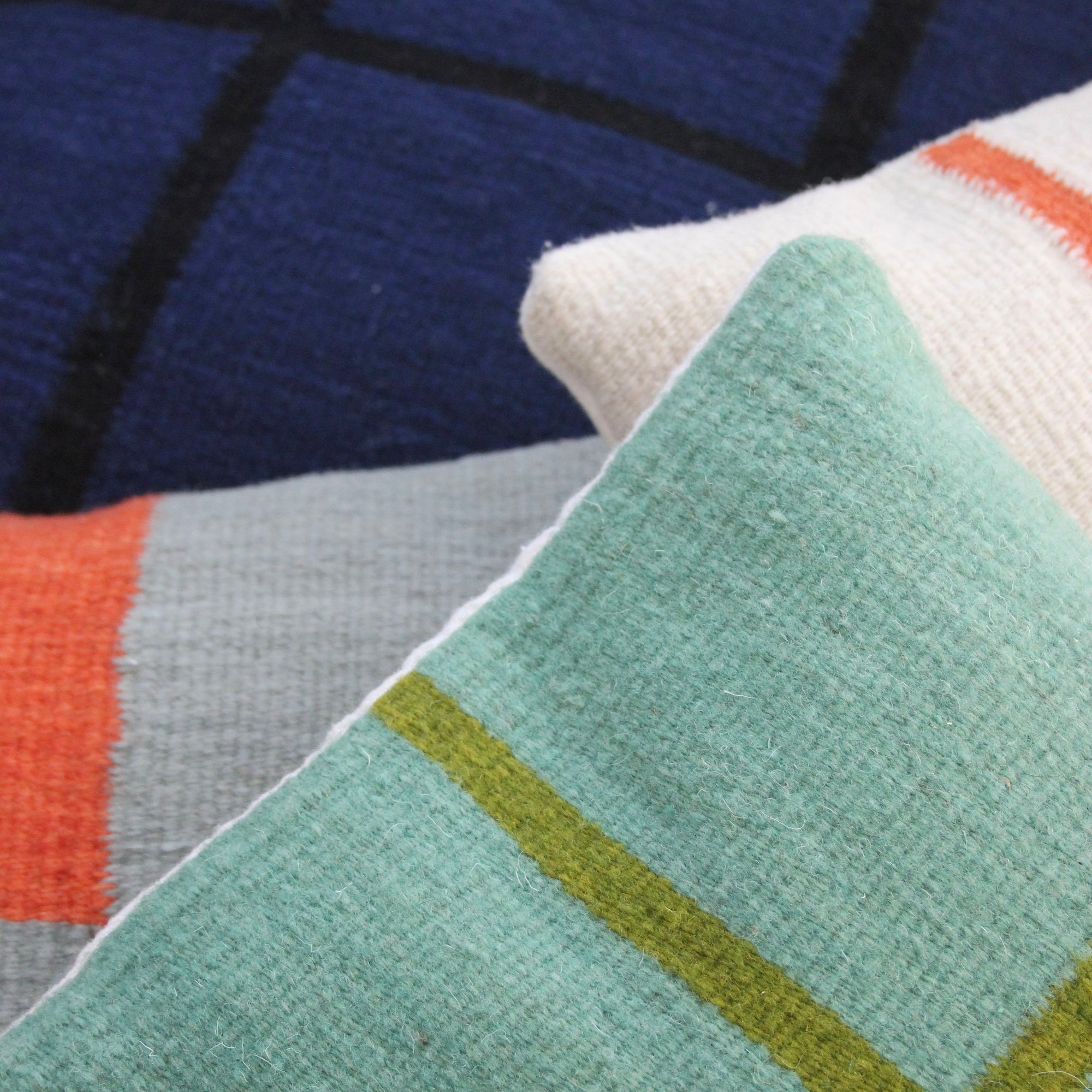 Three decorative pillows overlap: Ania Brzeski’s Pillow 07 in jade and olive stripes, a dark blue pillow with black modernist lines, and a cream pillow with orange and light blue stripes.