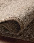 A close-up of the Loloi Alina Rug - Bark, a light brown hand-woven rug featuring a textured surface and subtle checkerboard design, partially rolled on a dark wooden floor.
