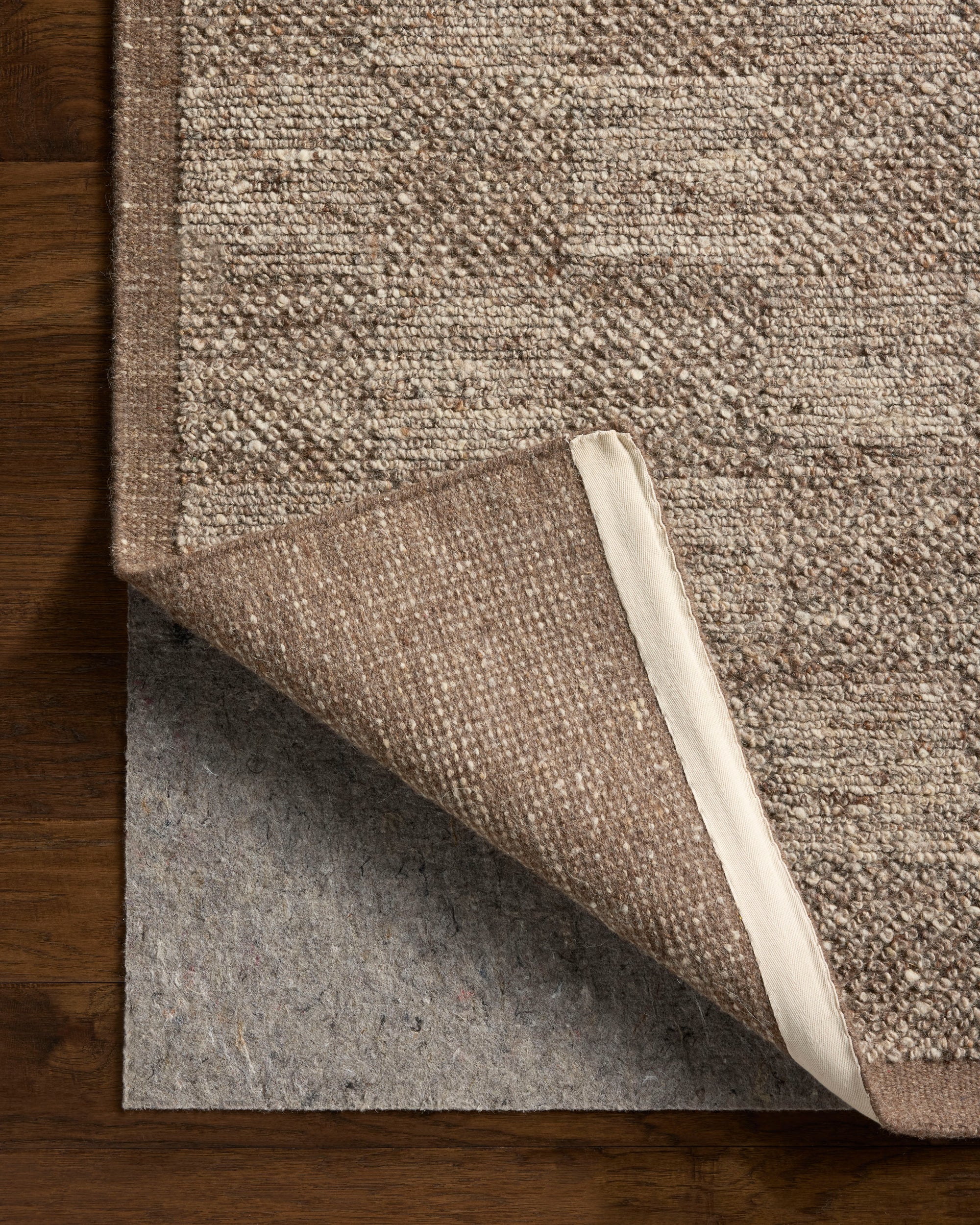 The Loloi Alina Rug - Bark, in shades of beige and taupe, is displayed on a wooden floor with one corner lifted to reveal a grey felt rug pad underneath.