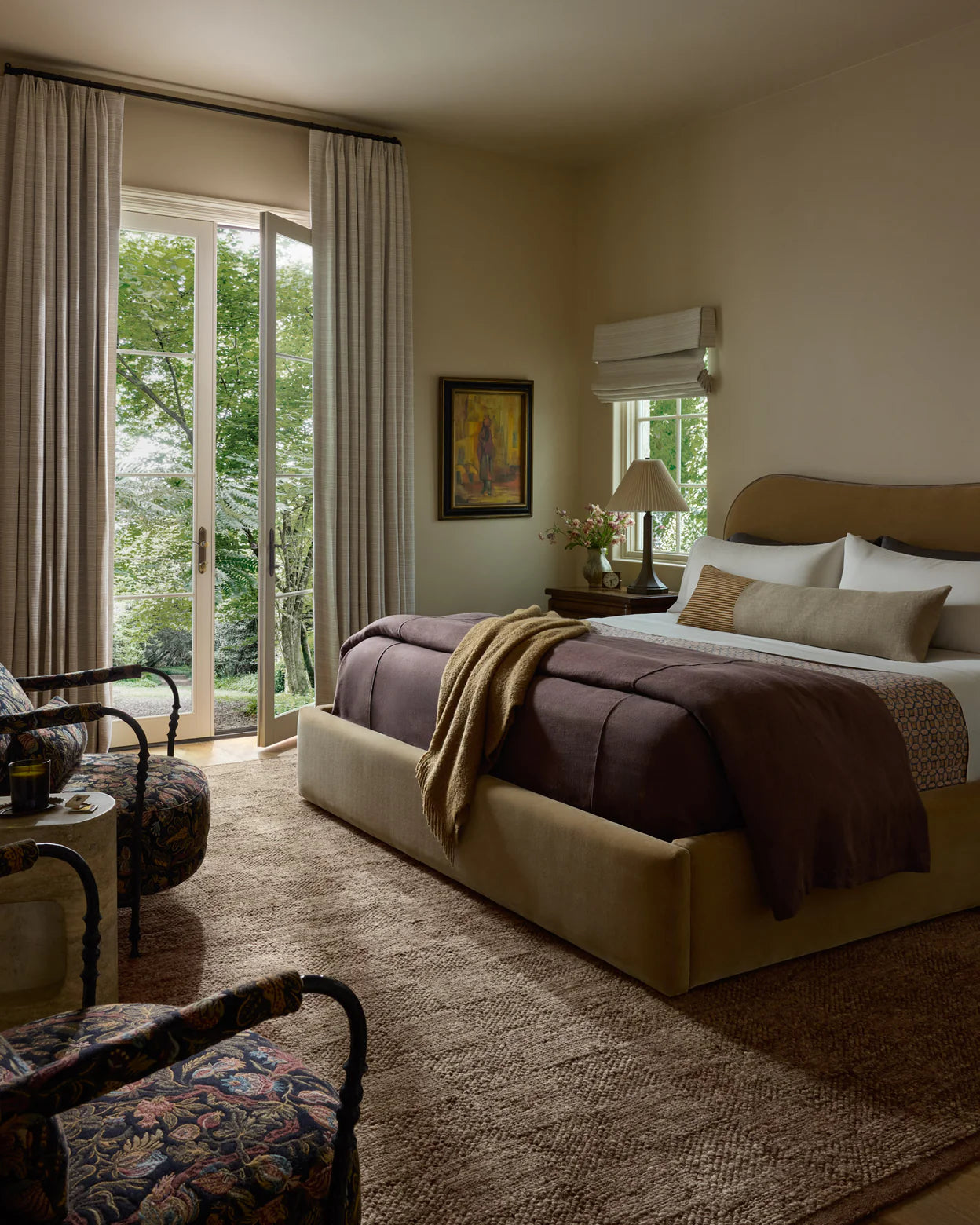 A cozy bedroom features a large bed with brown and beige linens, two patterned armchairs, and a window overlooking a garden. The Alina Rug - Bark by Loloi adds warmth, while soft lighting and neutral decor create an inviting atmosphere.