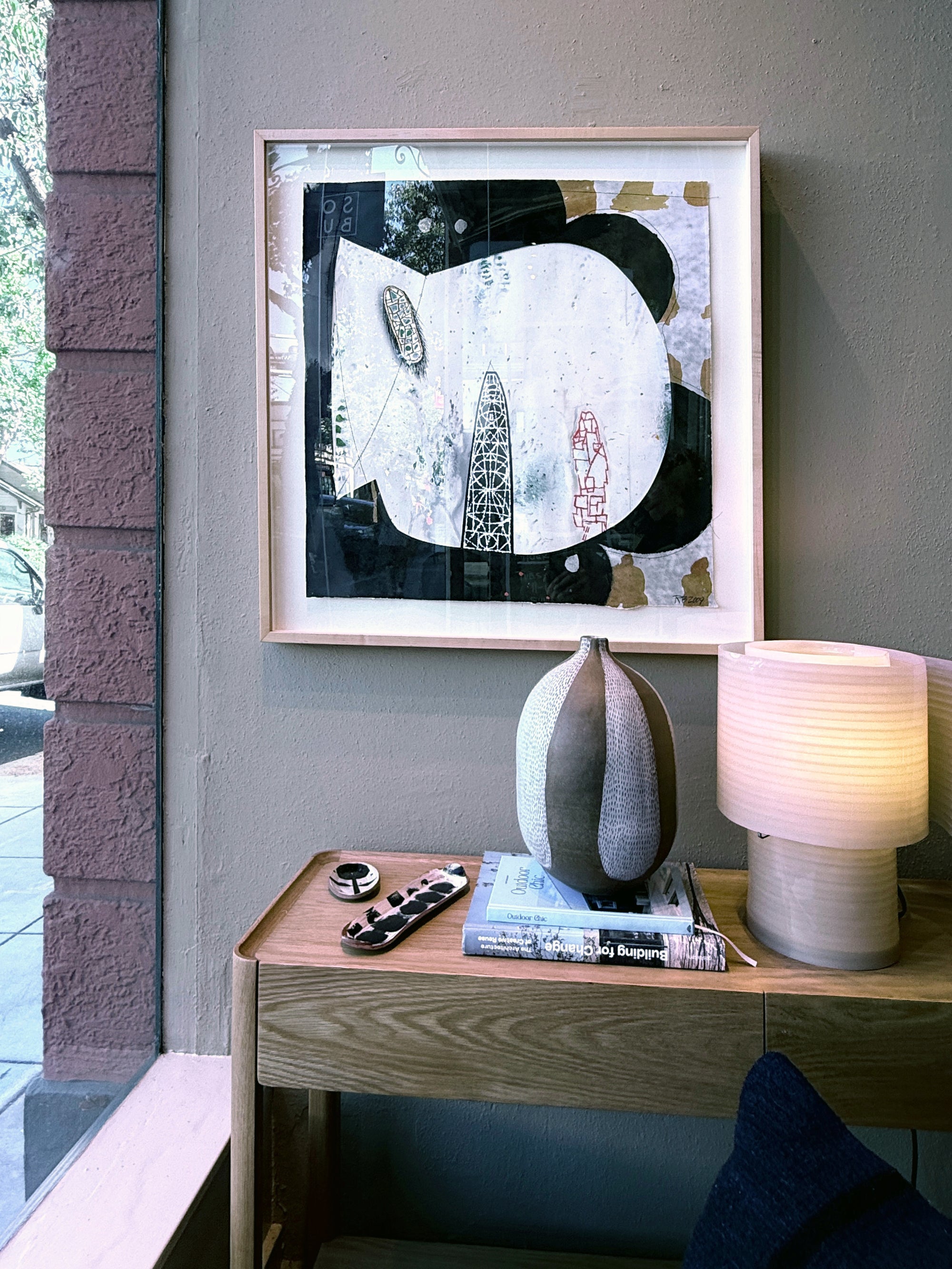 A wooden table by a window holds decor, while above it hangs "Area 51 Series" by ROBERT BRADY—a black and white abstract mixed media artwork in a wooden frame on a gray wall.