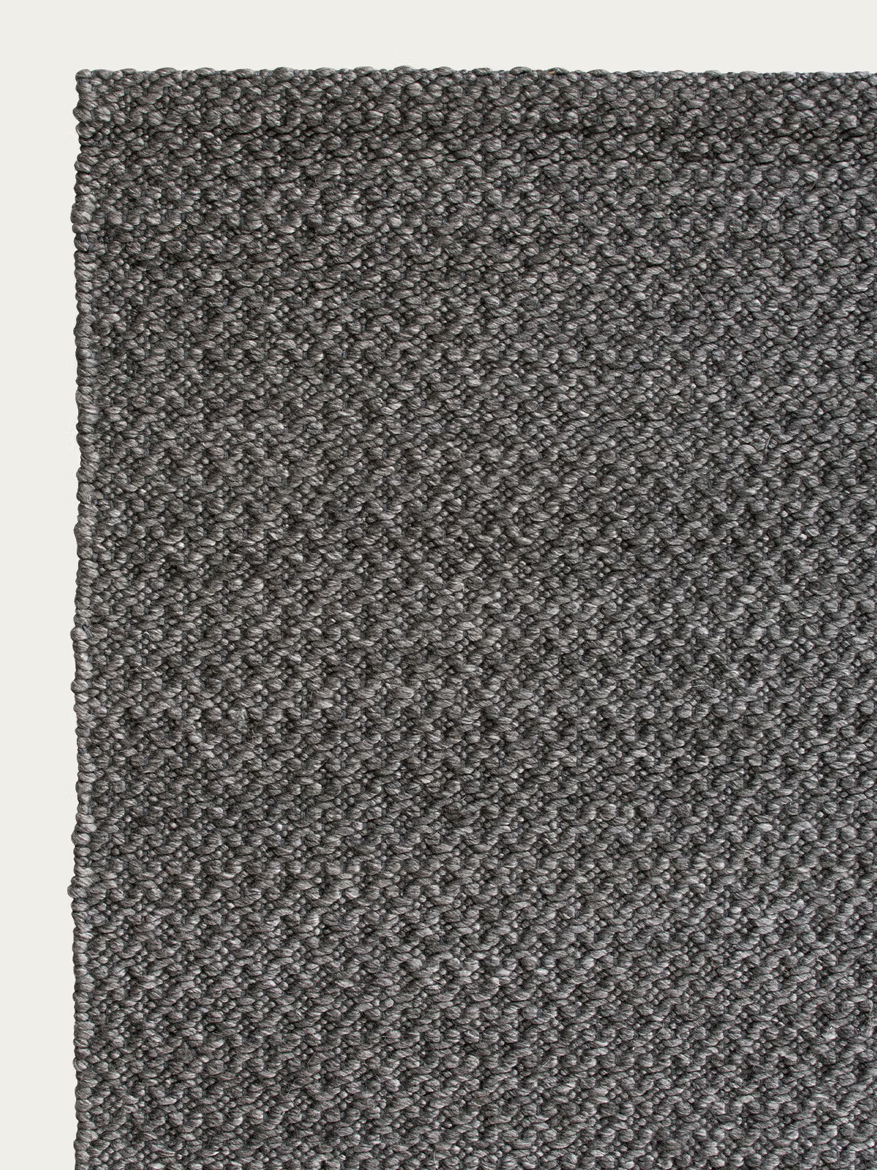 Close-up of the Sherpa Rug - Charcoal by Armadillo&amp;Co, featuring a dark gray, textured woven wool surface seen from above, with only the top left corner visible against an off-white background. This handwoven rug brings subtle depth and warmth to any space.