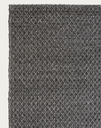Close-up of the Sherpa Rug - Charcoal by Armadillo&Co, featuring a dark gray, textured woven wool surface seen from above, with only the top left corner visible against an off-white background. This handwoven rug brings subtle depth and warmth to any space.