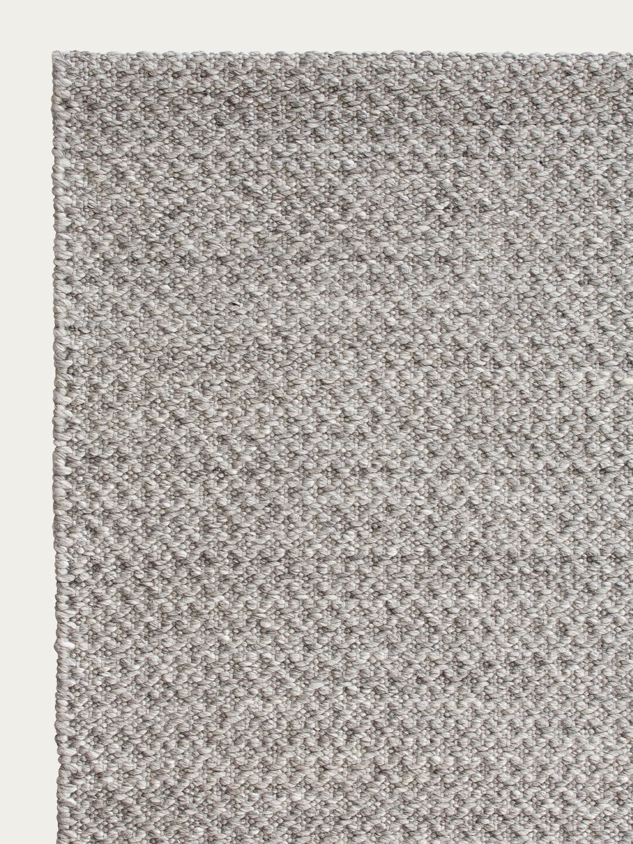 Close-up of the Sherpa Rug - Pumice by Armadillo&amp;Co, featuring a textured, light gray handwoven wool surface with a subtle diamond pattern, shown from the top left corner against an off-white background.