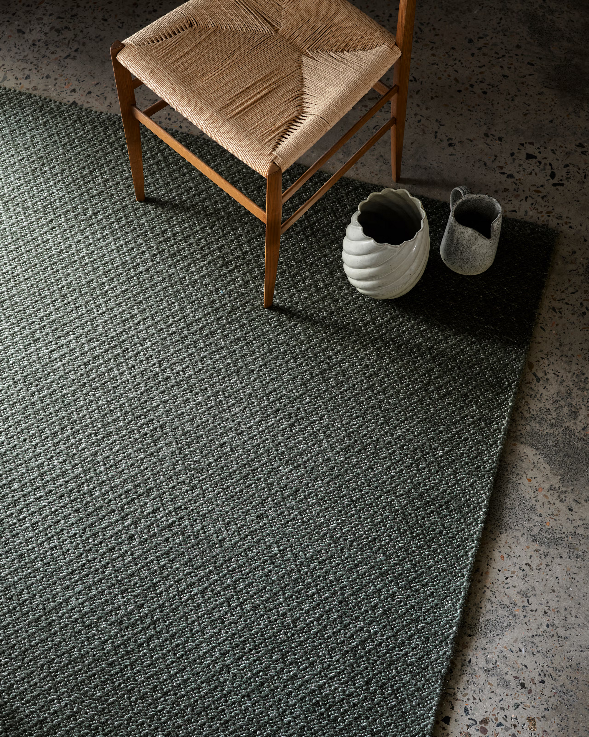 A woven chair sits on the Armadillo&Co Sherpa Rug in Kelp—a textured dark gray, hand-woven wool design—beside two decorative ceramic vases on a speckled concrete floor.