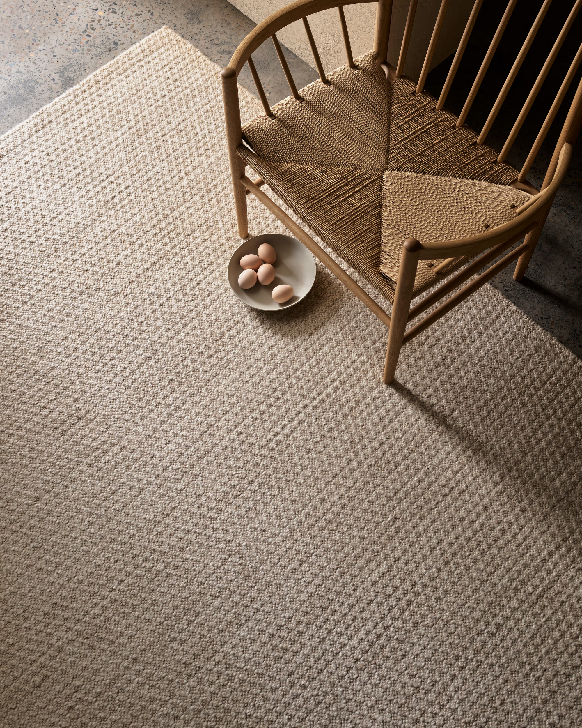 A wooden chair with a woven seat sits on the Sherpa Rug - Sand by Armadillo&amp;Co. Nearby, a bowl holding six brown eggs rests on the rug. Neutral tones create a calm, minimalist setting.