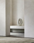 A minimalist space with textured off-white walls, a neutral stone sculpture with an oval cutout, and Armadillo&Co’s Odessa Rug in Caraway—an Afghan wool rug featuring natural yarn striations—creates a calm, serene setting.