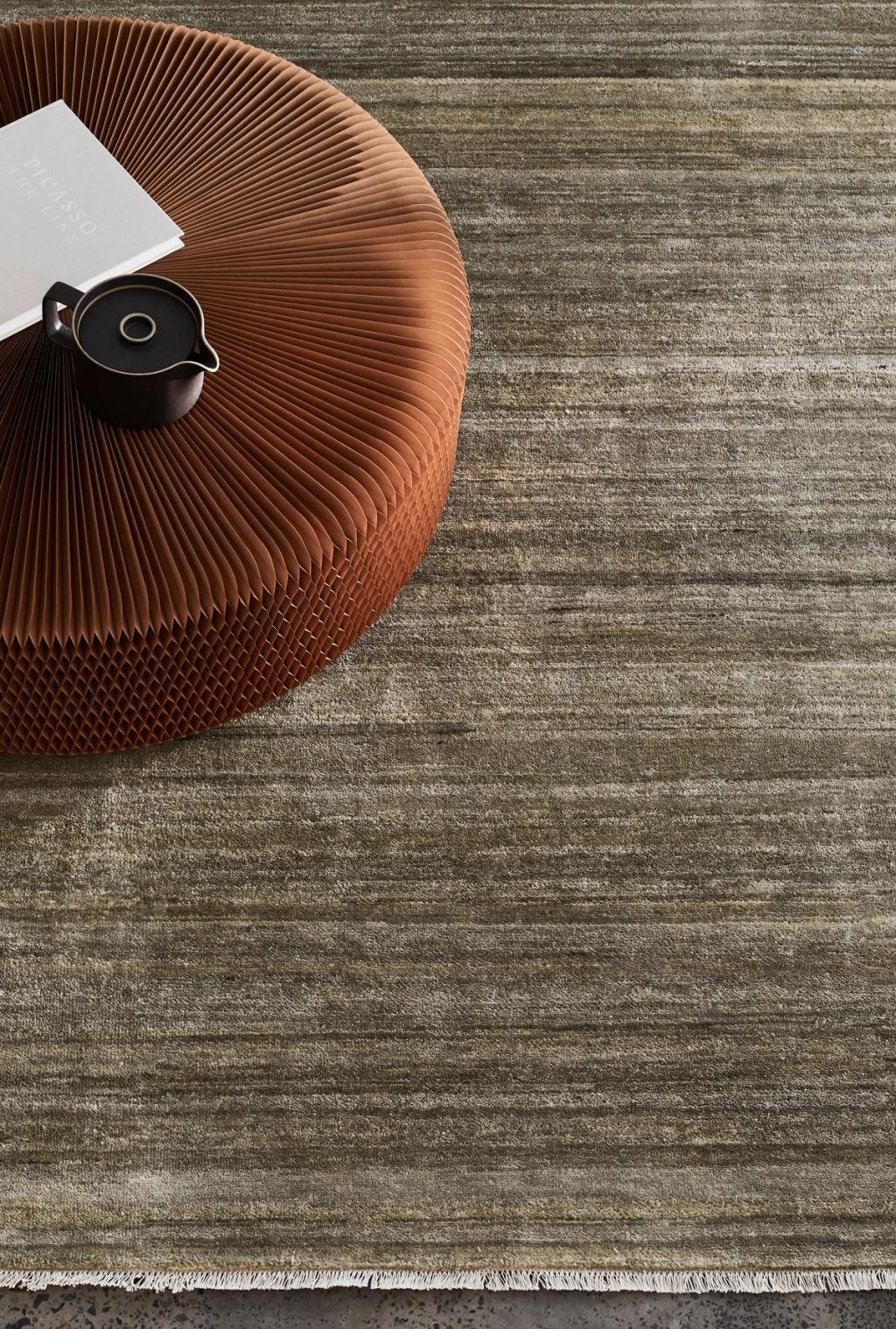 A textured brown ottoman with a white book and black teapot sits on the Armadillo&amp;Co Odessa Rug - Caraway, a muted greenish-gray Afghan wool rug with subtle horizontal patterns, natural yarn striations, and fringed edges.