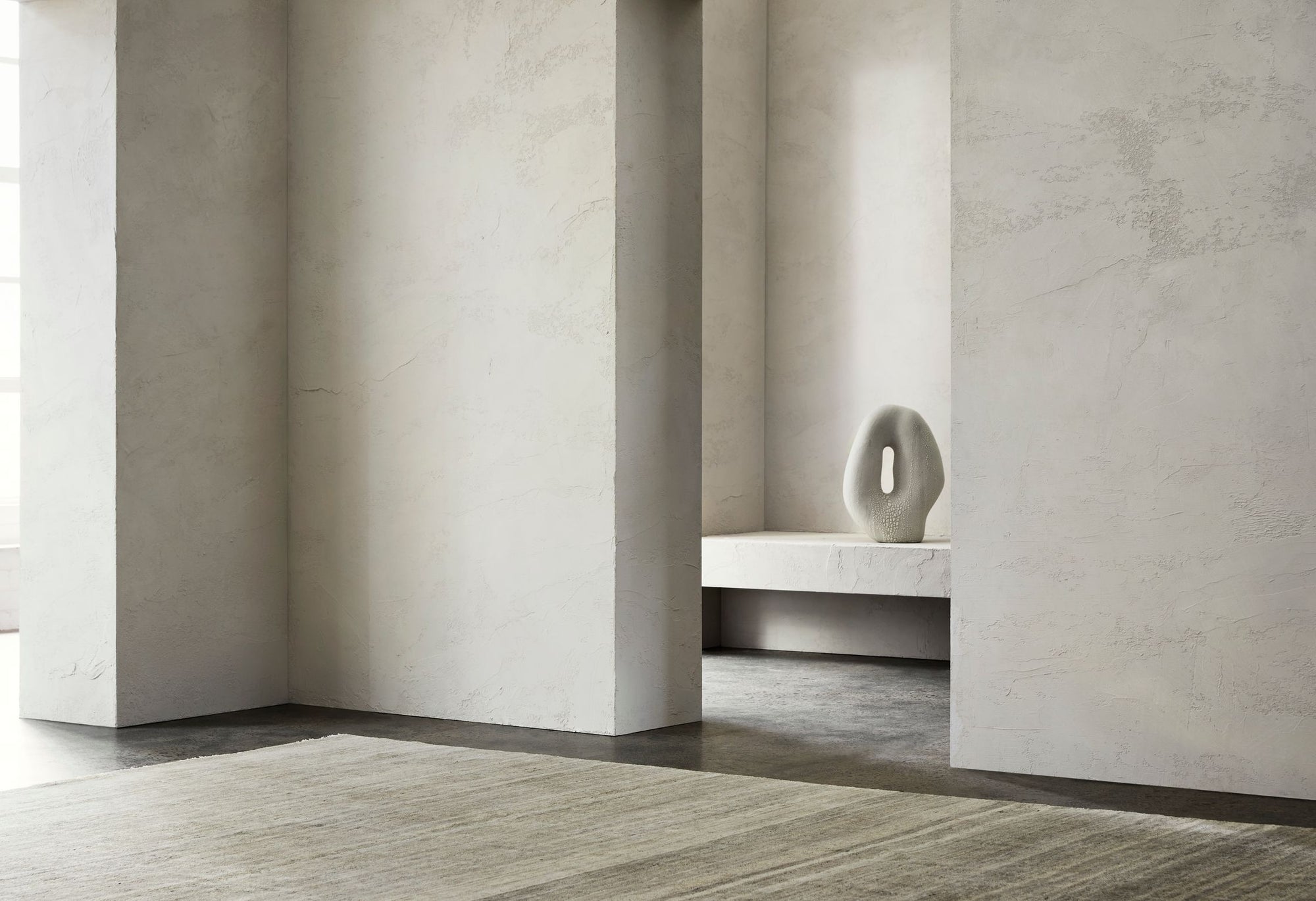 A minimalist interior features textured light gray walls, dark floors, and the Armadillo&Co Odessa Rug in Caraway. A white sculptural vase rests on a bench by the wall, illuminated by natural light filtering in from the left.